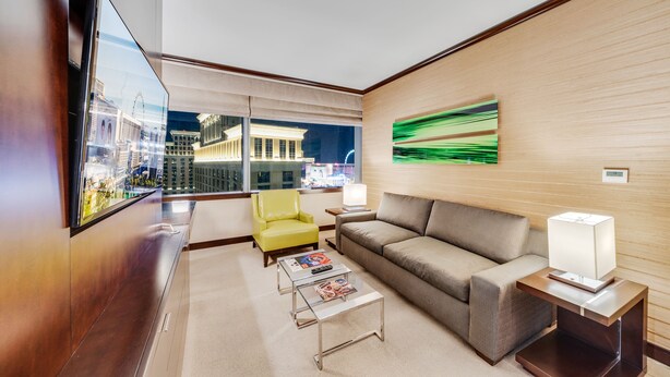 A modern hotel room with a large window offering a city view, a sofa, a yellow armchair, a coffee table, and a lamp. - Vdara Studio by Jet Luxury (Las Vegas)