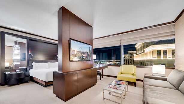 A modern hotel room with a large bed, a desk, a yellow armchair, a grey sofa, and a city view through large windows. - Vdara Adjoining Parlor Suite by Jet Luxury (Las Vegas)