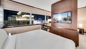 A hotel room with a large bed, a TV mounted on a wooden cabinet, a desk with a chair, and a view of a cityscape including the Eiffel Tower. - Vdara Adjoining Parlor Suite by Jet Luxury (Las Vegas)