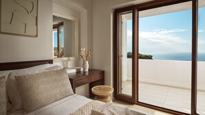 Een slaapkamer met een bed, nachtkastje, vaas met gedroogde bloemen, houten kruk en een grote schuifdeur naar een balkon met uitzicht op zee.