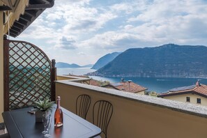 A balcony with a table, chairs, and a bottle of wine. The view includes a lake and mountains in the distance.