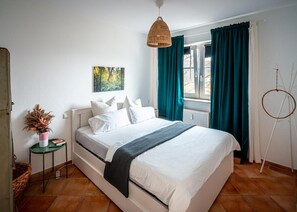 Un lit bien fait, avec du linge de lit blanc et une couverture sombre, un luminaire suspendu en osier, un tableau représentant une forêt et des rideaux turquoise à une fenêtre.