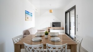 A modern dining area with a wooden table set for four, white chairs, a sofa, and a TV in the background. - Homiday - Waterfront, Interior 9 (Borgo Santa Maria Immacolata)