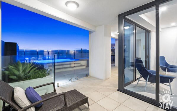 A modern balcony with a glass railing, a pool, and a seating area with chairs.