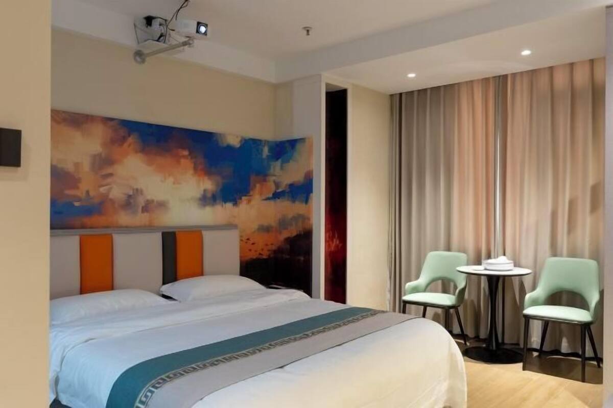 Habitación de hotel con una cama grande, un proyector, dos sillas y una pequeña mesa.