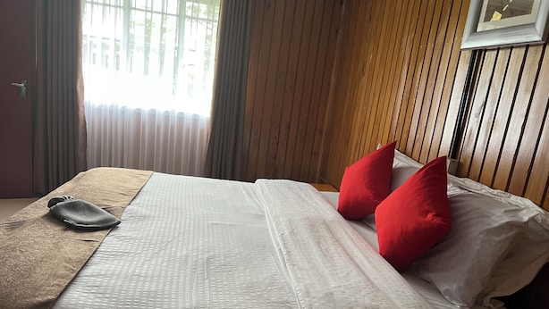 Una cama bien hecha con sábanas blancas y dos almohadas rojas, un plaid marrón y un cabecero de madera.