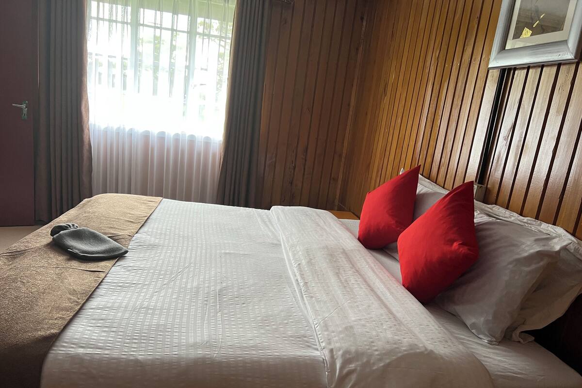 A neatly made bed with white linens and two red pillows, a brown throw blanket, and a wooden headboard.