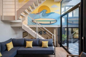 Ein modernes Interieur mit Treppe, Wandgemälde einer Welle und einem dunkelgrauen Sofa mit gelben Kissen.