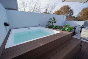 Ein modernes Außen-Whirlpool mit Holzterrasse, umgeben von Grün und einer weißen Wand.