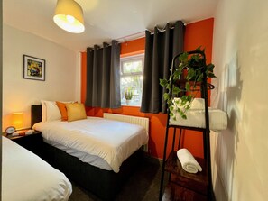A hotel room with two beds, a window with curtains, a radiator, a bedside lamp, a clock, a framed picture on the wall, and a plant on a shelf. - Curzon House 1 (Cleethorpes)