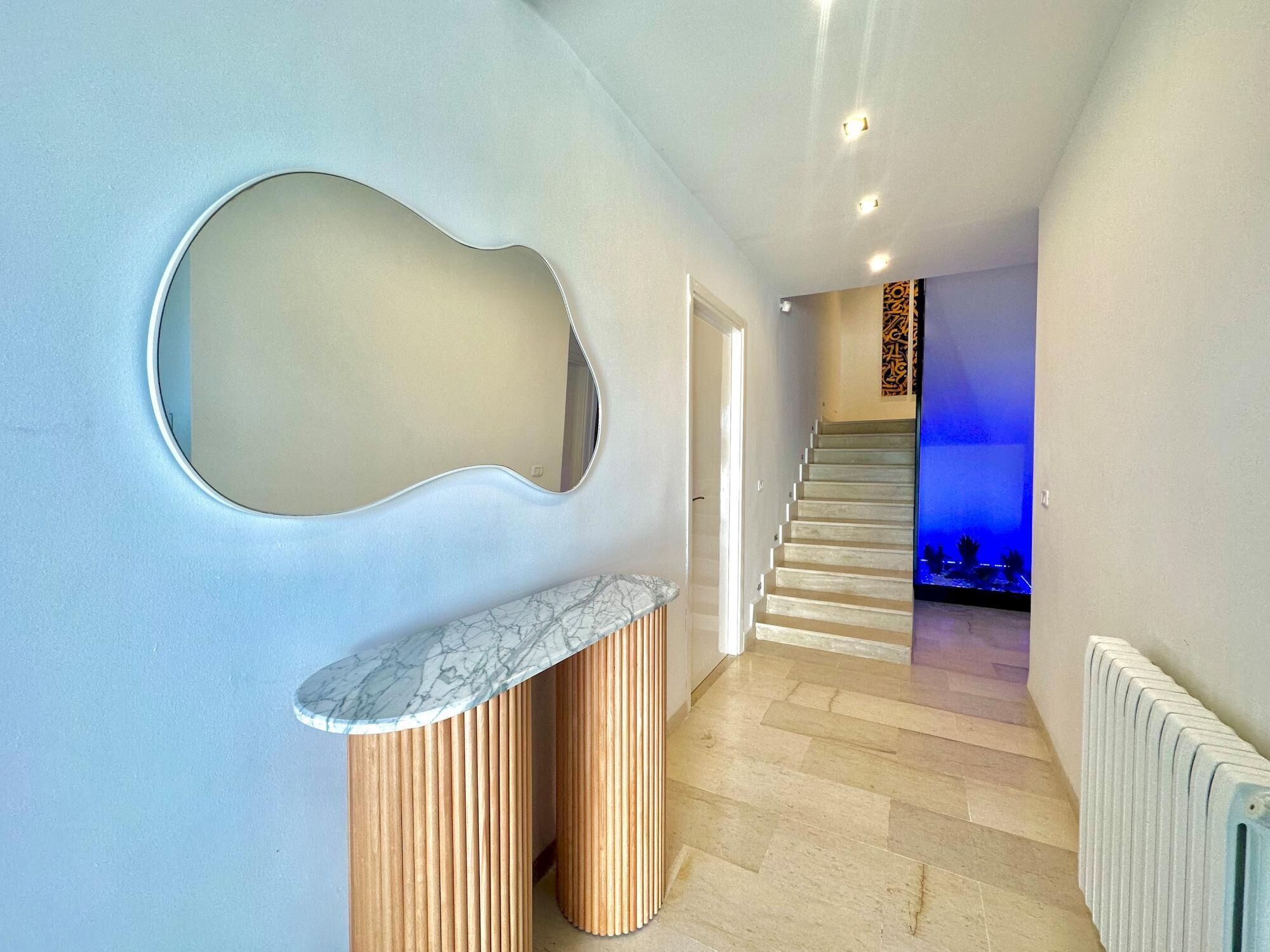 A modern interior with a curved mirror, a marble countertop, wooden slatted columns, a staircase, and a blue-lit room in the background.
