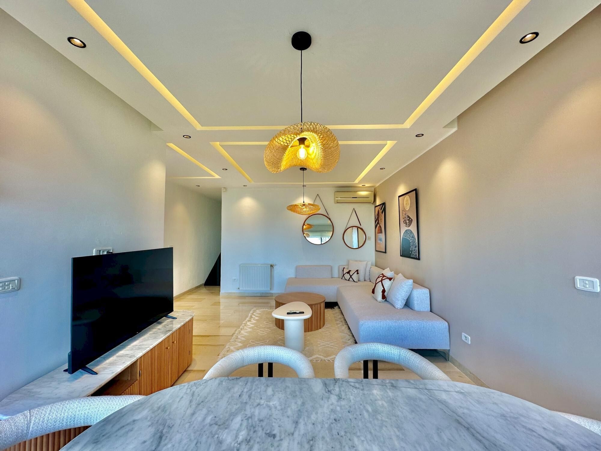 A modern living room with a flat-screen TV, a marble dining table, a sofa, and a hanging light fixture.
