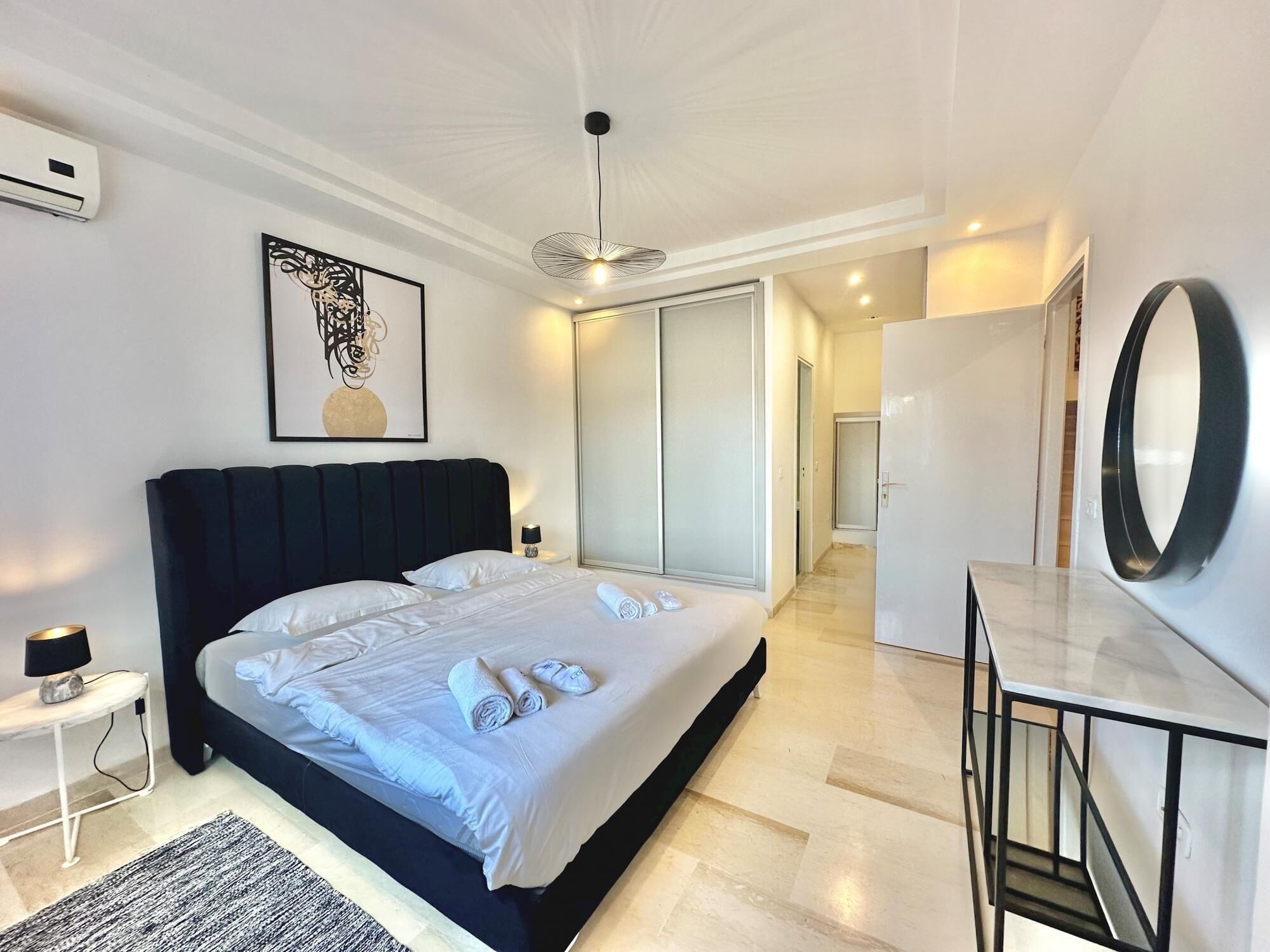 Una habitación de hotel moderna con una cama grande, una mesita de noche con lámpara, un espejo y una obra de arte enmarcada en la pared.