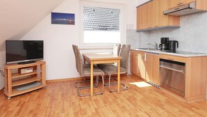 A compact kitchen with wooden cabinets, a built-in oven, and a dining area with a TV and a small table. - Haus Wirdemann Wohnung 2 (Norderney)