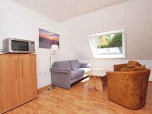 Una sala moderna con un sofá, una mesita, un microondas sobre un mueble, una lámpara de pie y una ventana con vistas a la vegetación.