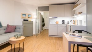 Ein modernes, kompaktes Wohnzimmer mit grauem Sofa, einem kleinen runden Tisch, einer Küchenzeile mit weißen Schränken und einer Essecke mit Holztisch und Stühlen.