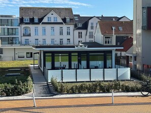 A modern building with a glass facade, a small outdoor seating area, and a paved pathway leading to it. - Haus Großer Seekaiser (Norderney)