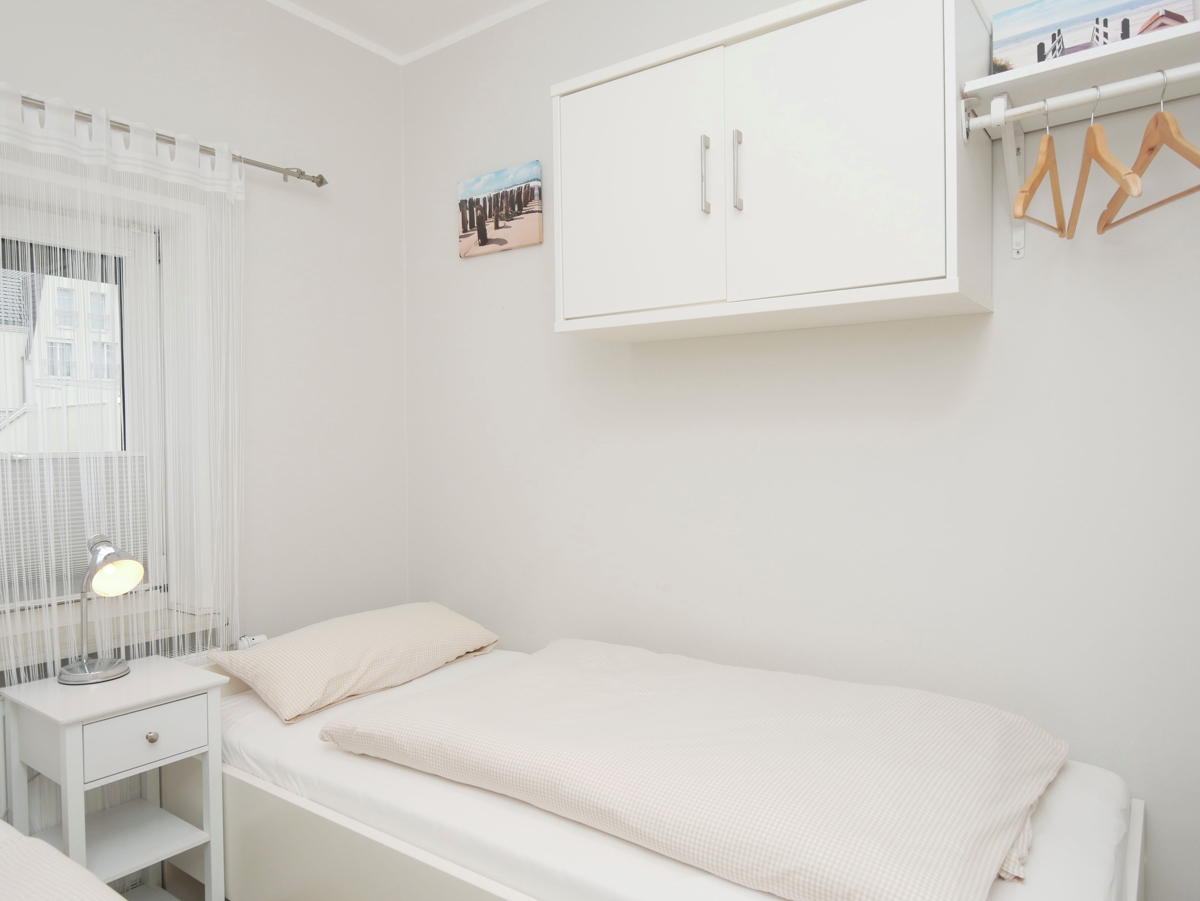 A bedroom with a single bed, a nightstand, a wardrobe, and a window with sheer curtains.