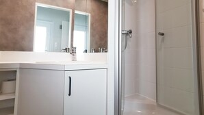 Une salle de bain moderne avec une douche à l’italienne en verre, un lavabo blanc avec un grand miroir et un meuble blanc doté d’une poignée noire.