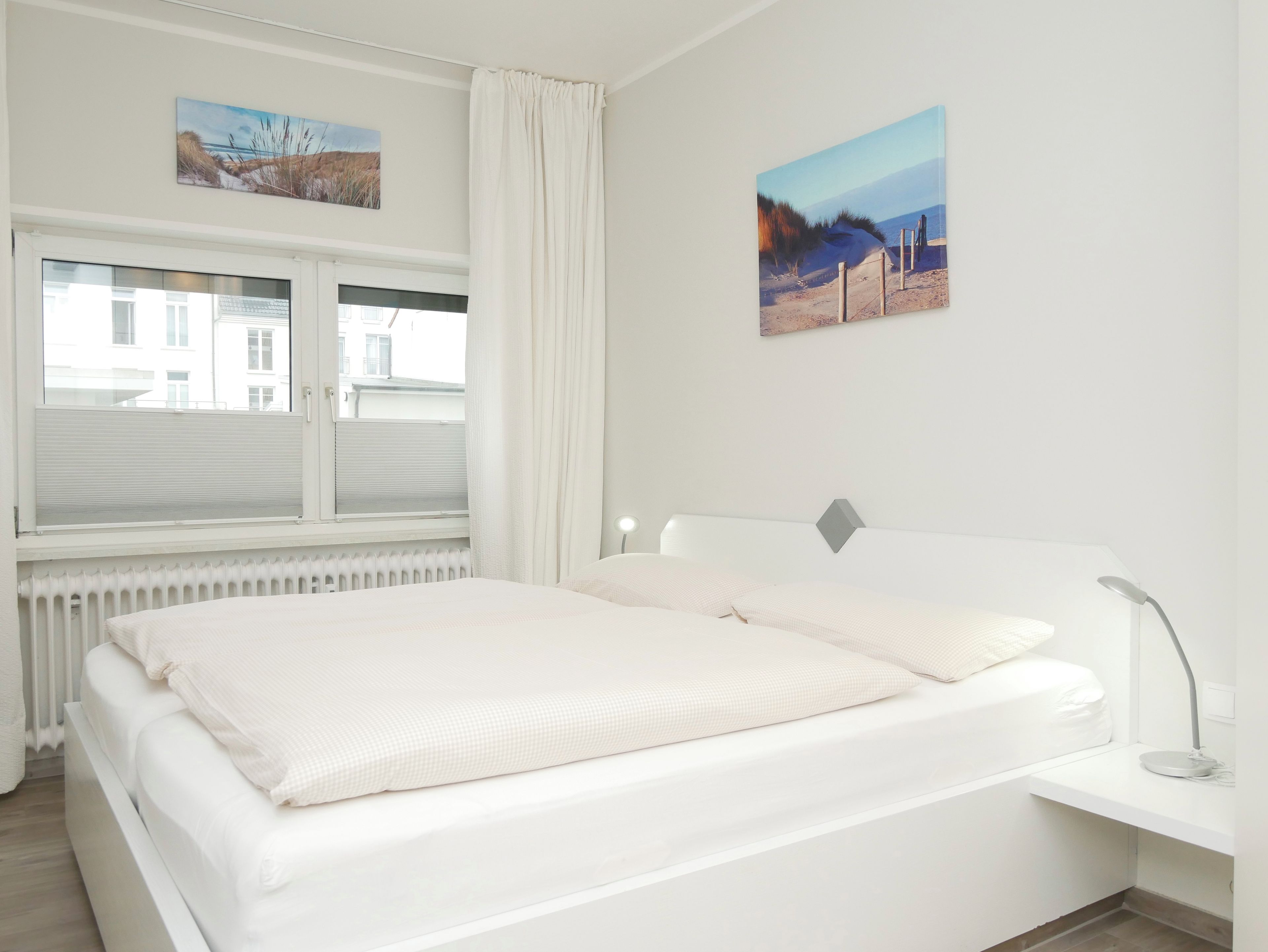 Quarto com cama, janela com cortinas, radiador e duas quadros pendurados na parede.