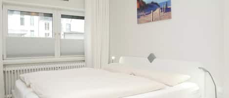 Quarto com cama, janela com cortinas, radiador e duas quadros pendurados na parede.