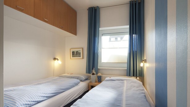 A small, compact room with two single beds, a wooden cabinet, a small table with a lamp, and a window with curtains. - House Seekaiser Little Seekaiser (Norderney)