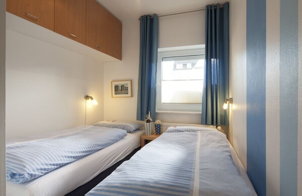 A small, compact room with two single beds, a wooden cabinet, a small table with a lamp, and a window with curtains. - House Seekaiser Little Seekaiser (Norderney)