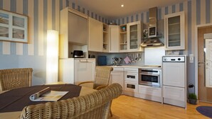 A compact kitchen with white cabinets, a built-in oven, and a microwave. There is a dining area with wicker chairs and a table. - House Seekaiser Little Seekaiser (Norderney)