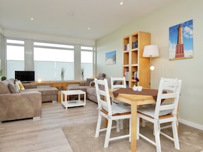 A modern living room with a sofa set, a dining table with chairs, a bookshelf, and a television. - Seekaiser House Little Seekaiser (Norderney)