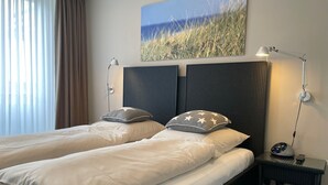 Quarto de hotel com duas camas, uma mesa de cabeceira com abajur, um quadro com grama e vista para o exterior.
