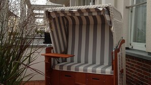 Uma cadeira de exterior com padrão listrado, uma mesa de madeira, um pequeno armário e uma janela com vista para a rua.