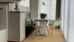 Uma cozinha moderna com fogão embutido, área de refeições com duas cadeiras e uma mesa, e uma janela grande com cortinas leves.