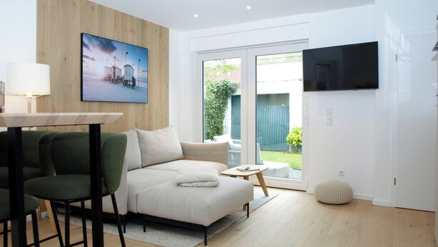 A modern living room with a sofa, a small table, a TV, and a view of the outdoors. - Wohnung Nr. 3 / Rübsamen-lüttenberg (Norderney)