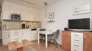 A modern kitchen with a microwave, coffee maker, and sink. There is a dining table with chairs, a TV on a wooden cabinet, and a small shelf with decorative items. - Haus Ulrichs Wohnung 1 b (Norderney)
