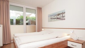 A bedroom with a bed, a nightstand, a lamp, a window with curtains, and a wall-mounted picture. - Haus Ulrichs Wohnung 1 b (Norderney)