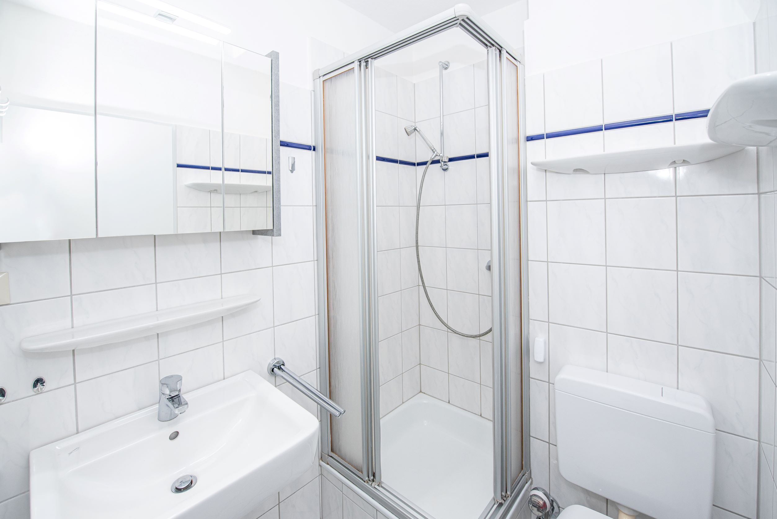 Un baño moderno con ducha envuelta en vidrio, un lavabo blanco con espejo arriba y un inodoro con un detalle azul.