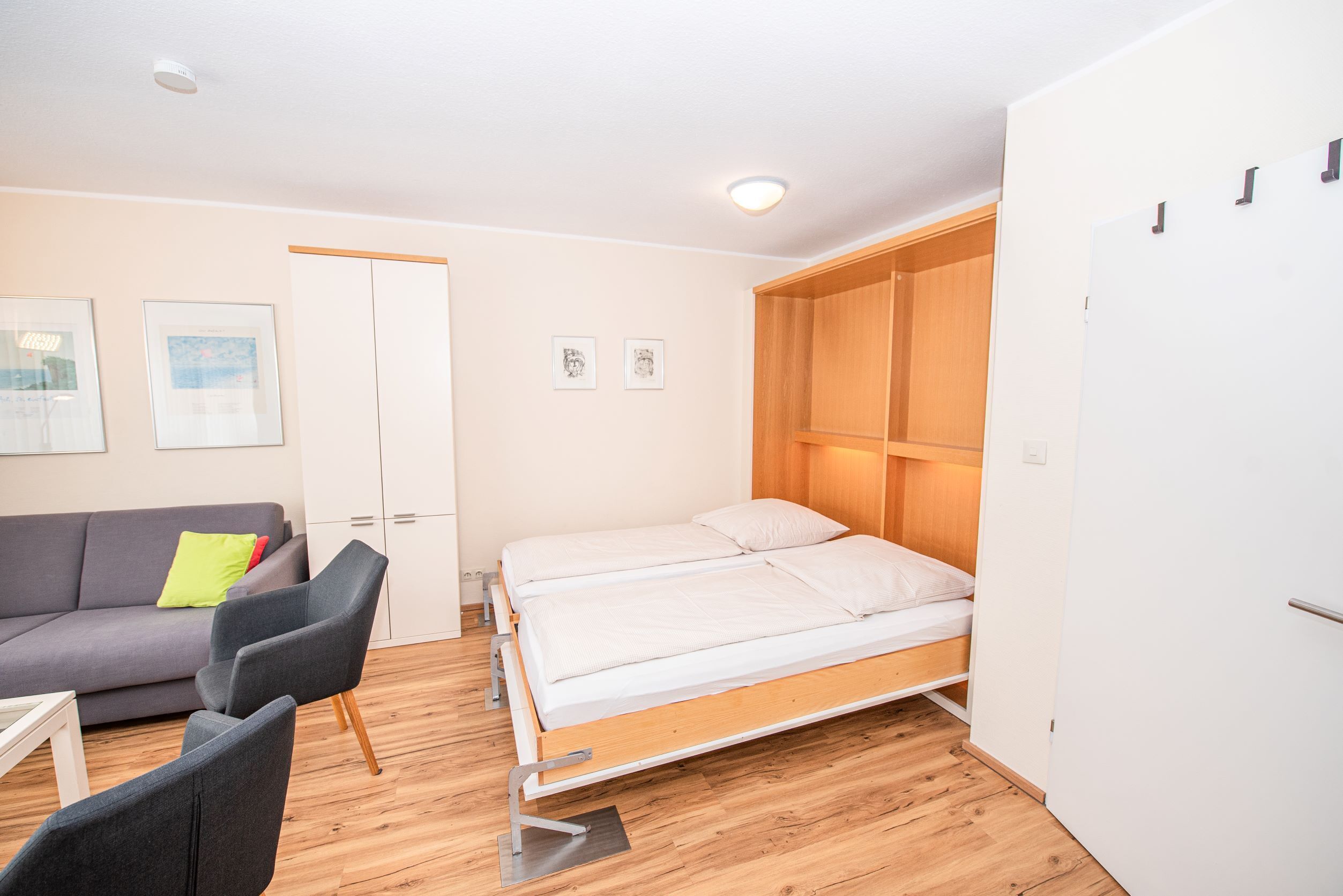 Una habitación compacta con una cama, un sofá gris con un cojín verde, una mesita y un armario.