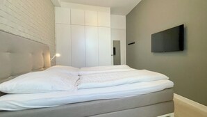 A modern bedroom with a large bed, a white headboard, a built-in wardrobe, and a flat-screen TV mounted on the wall. - Haus Jantje Mö Wohnung 5 (Norderney)