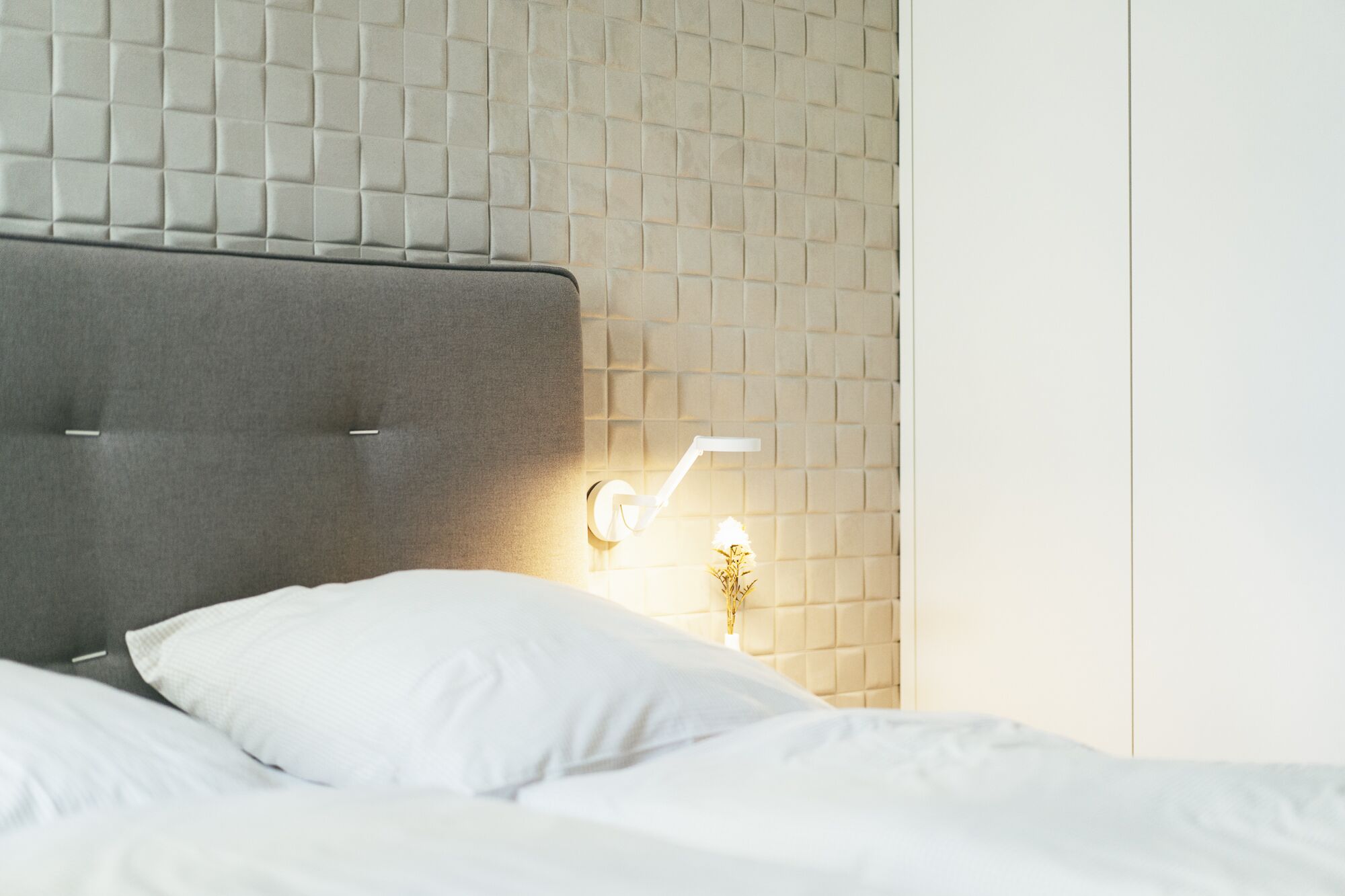 Ein Bett mit grauem Kopfteil, einem weißen Kopfkissen und einer Wandleuchte mit einer kleinen Pflanze.