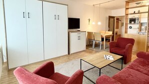 En moderne stue med røde polstrede stole, et træbord og et fjernsyn. Der er et spiseområde med bord og stole, og et køkken i baggrunden.