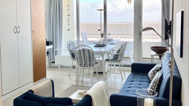 A modern living room with a sofa, chairs, a dining table, and a view of the beach. - Haus Meeresbrandung Wohnung 23 (Norderney)