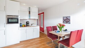 Une cuisine moderne avec des meubles blancs, un four encastré et une machine à café. On y trouve également un coin repas avec une table en bois et des chaises rouges.