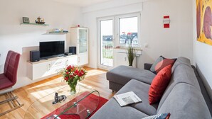 Un salon moderne avec un canapé gris, des coussins rouges, une table basse en verre et un téléviseur à écran plat posé sur un meuble blanc.