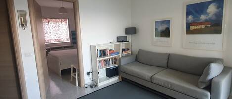 A living room with a sofa, a bookshelf, a desk, and a window with patterned curtains.