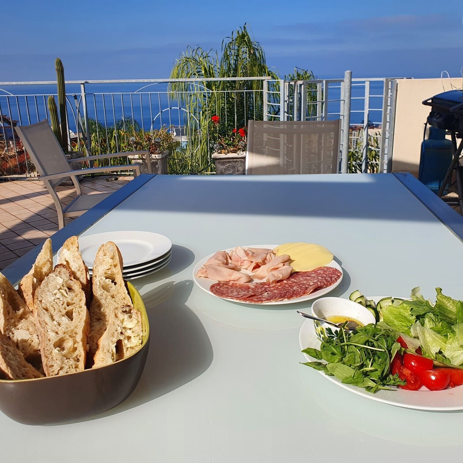 An outdoor dining table with a variety of food items including bread, cured meats, cheese, and a salad, set against a backdrop of a clear blue sky and a view of the sea.