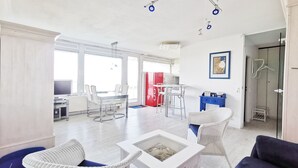 Una sala de estar moderna con una mesa de centro de vidrio, sillas de mimbre blanco, un sofá azul y una zona de cocina con un refrigerador rojo.