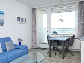 Een moderne woonkamer met een blauwe bank, een glazen salontafel, een eettafel met stoelen, een boekenkast en een groot raam met uitzicht op zee.