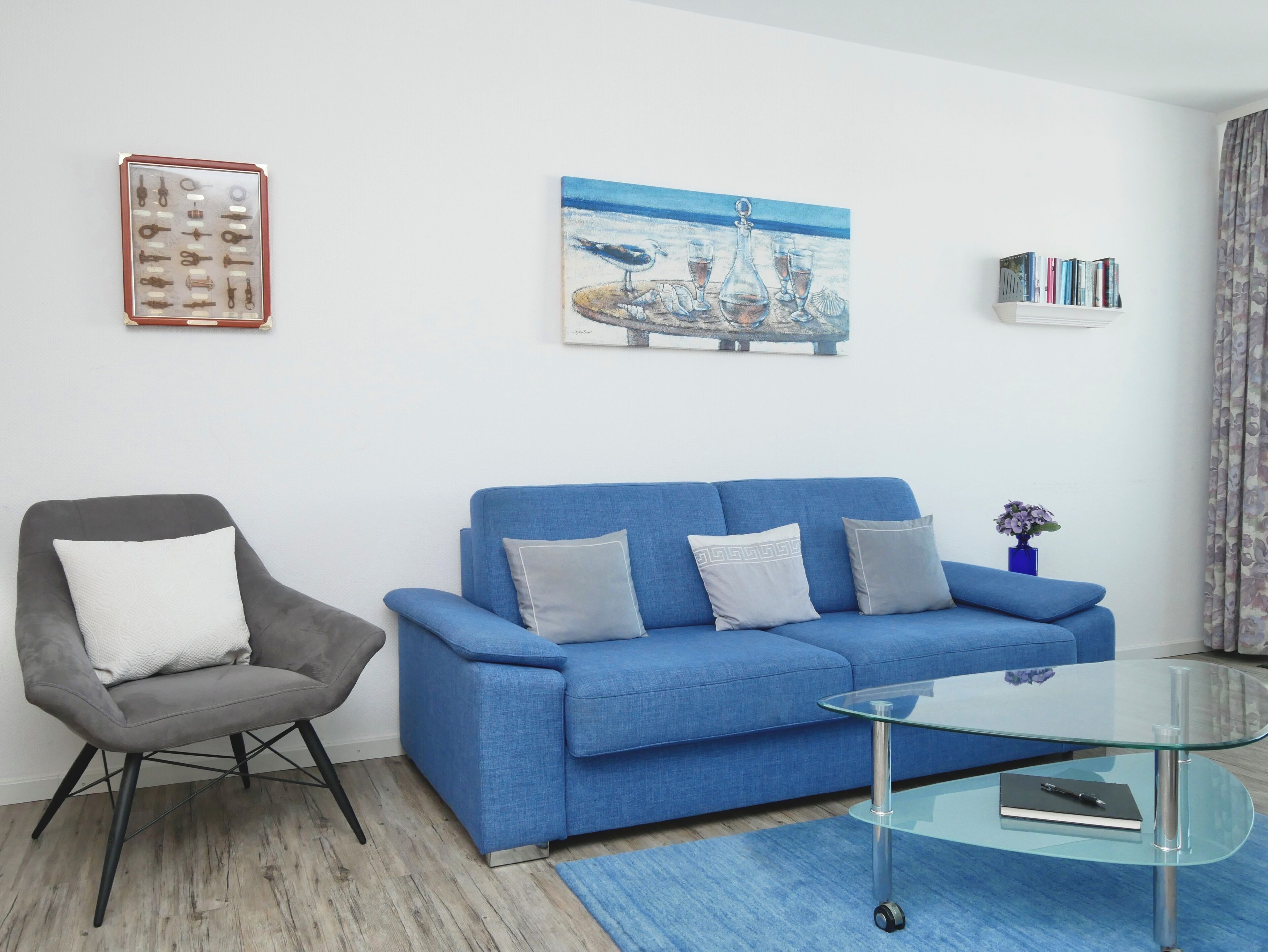 A living room with a blue sofa, a grey armchair, a glass coffee table, a painting, and a bookshelf.