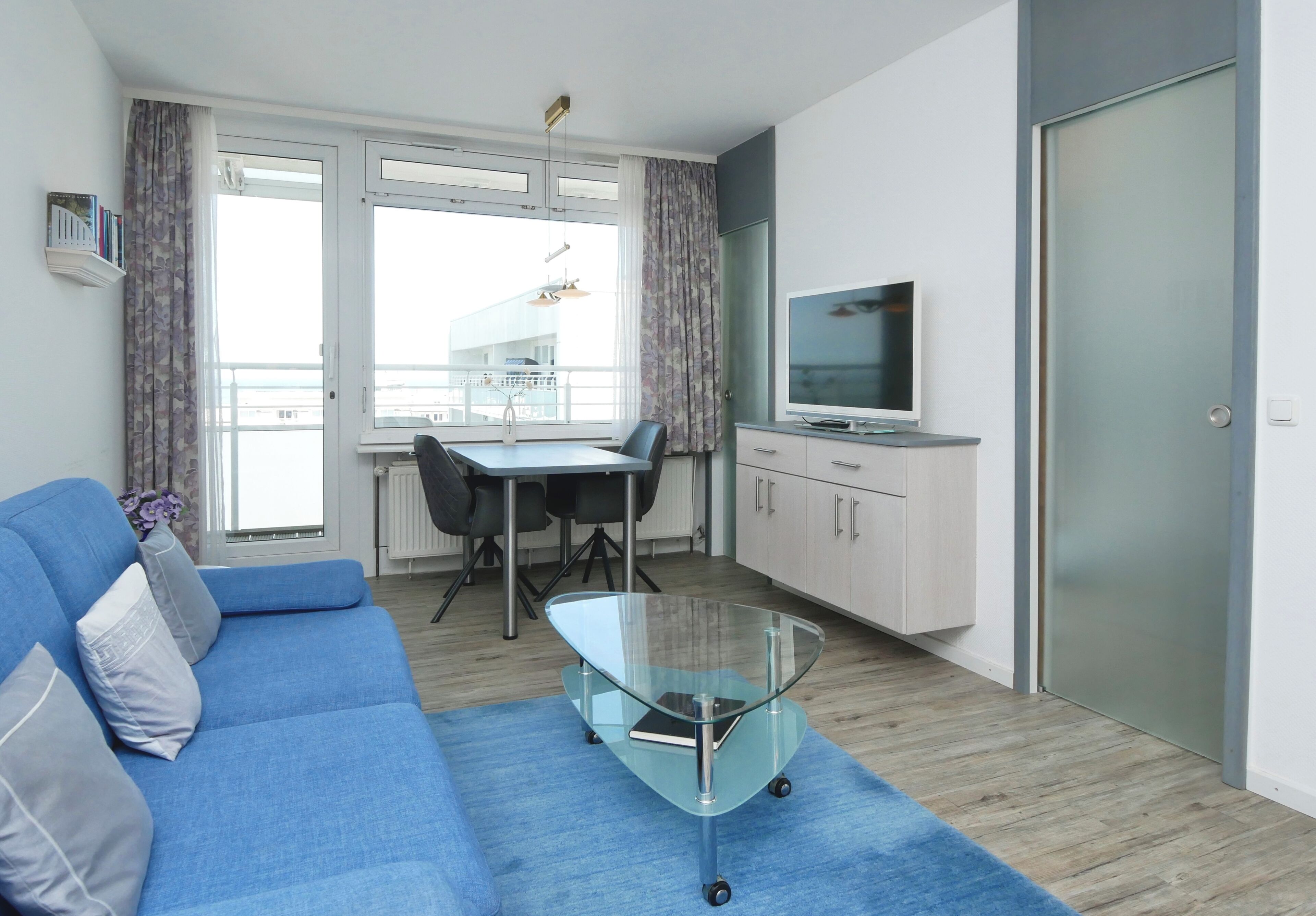Uma sala de estar moderna com um sofá azul, uma mesa de centro de vidro, uma televisão e um espaço de refeições com mesa e cadeiras.
