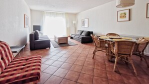 A living room with a sofa, a dining table with chairs, a TV, and a window with curtains. - Villa Nordland Wohnung 6 (Norderney)